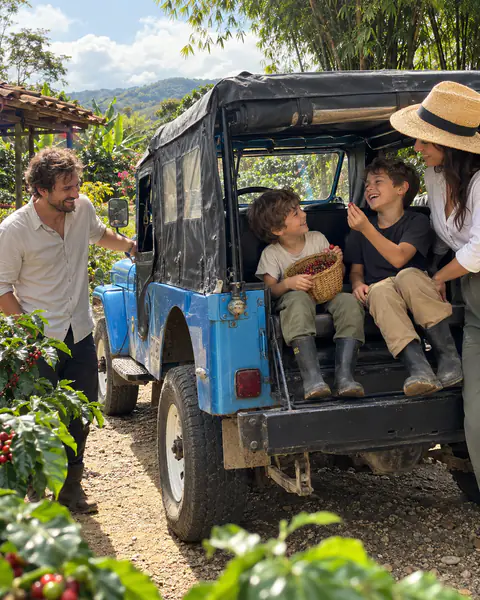 Eine Familie neben einem blauen Willys in einer Kaffeeplantage, die Kinder halten einen Korb mit Kaffeekirschen
