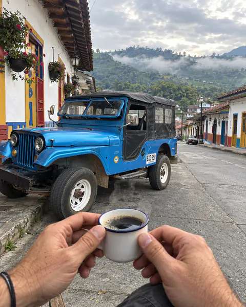 Ein tinto in der Hand vor dem blauen Willys, in einer bunten Straße im Quindío